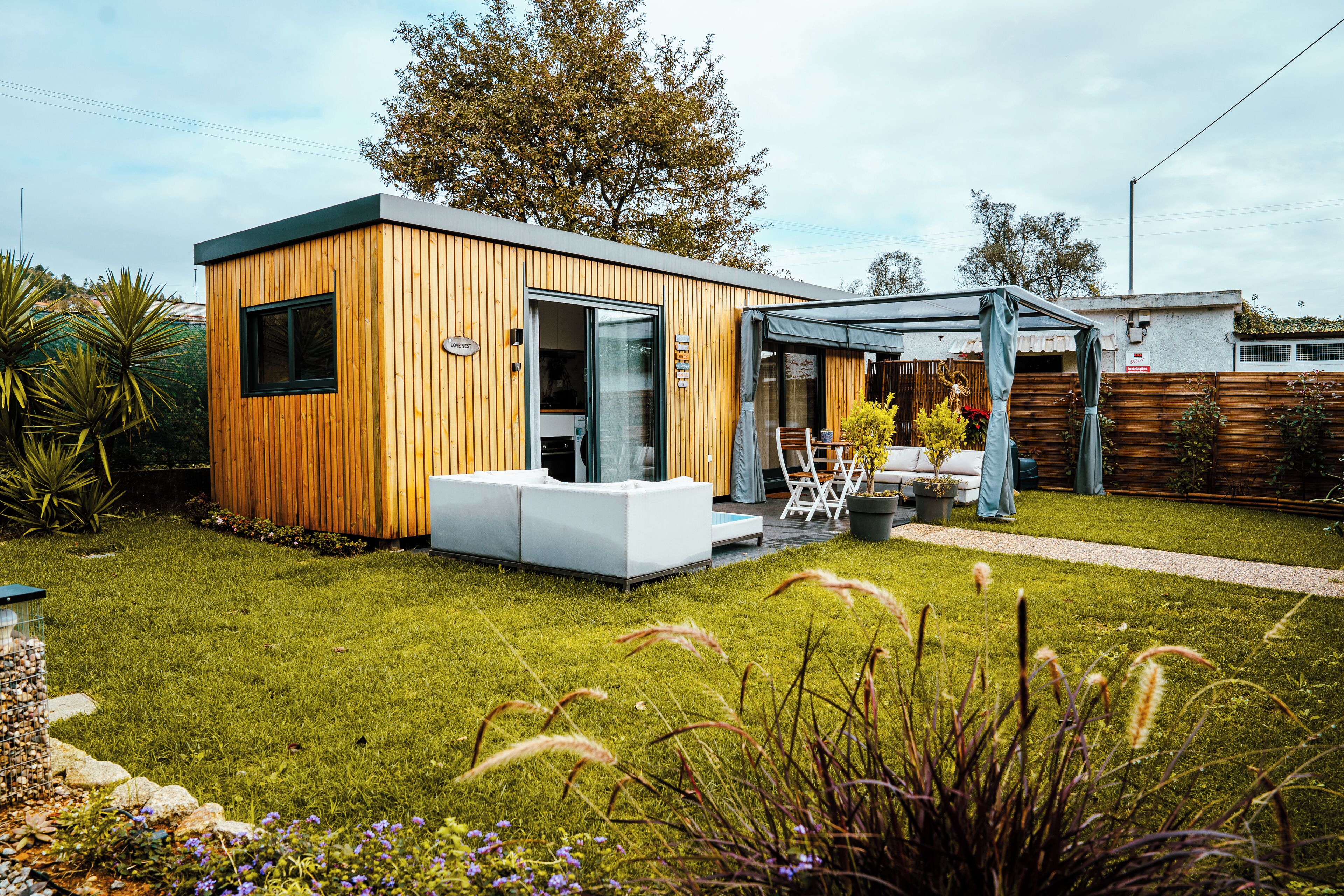 Une maison moderne en bois, dotée d’une porte en verre, d’un espace extérieur aménagé et couvert, et d’une pelouse bien entretenue.