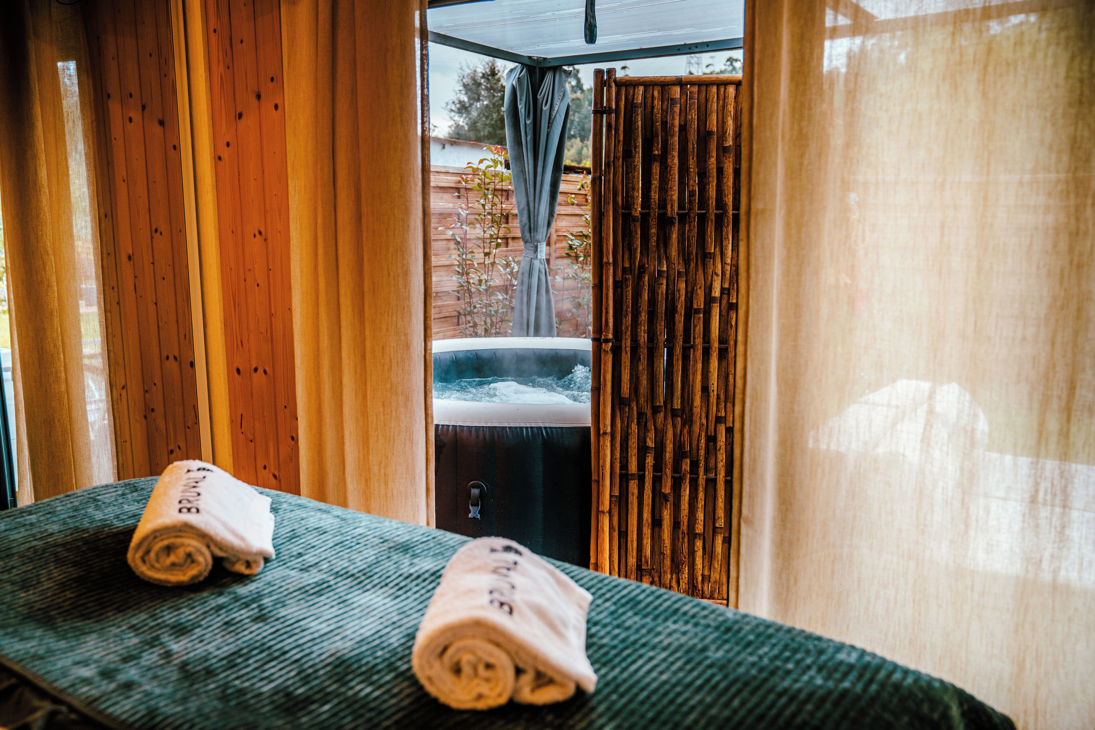 Ein Wellnessraum mit Holzwand, Massagetisch und einer sichtbaren Whirlwanne hinter einem Bambusvorhang.