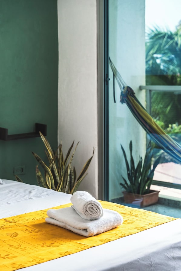 A hotel room with a bed, a yellow patterned bedspread, a rolled white towel, and a view of a hammock and plants outside the window. - Villa Nuevo Amanecer Santa Verónica (Juan de Acosta)