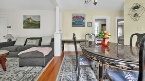 Ein Wohnzimmer mit Sofa, Hocker und einem Esstisch mit Stühlen. An den Wänden hängen Bilder und auf dem Tisch steht eine Vase mit Blumen.