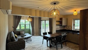 A spacious living area with a sofa, dining table, and chairs, a kitchen with a modern design, and large windows allowing natural light.