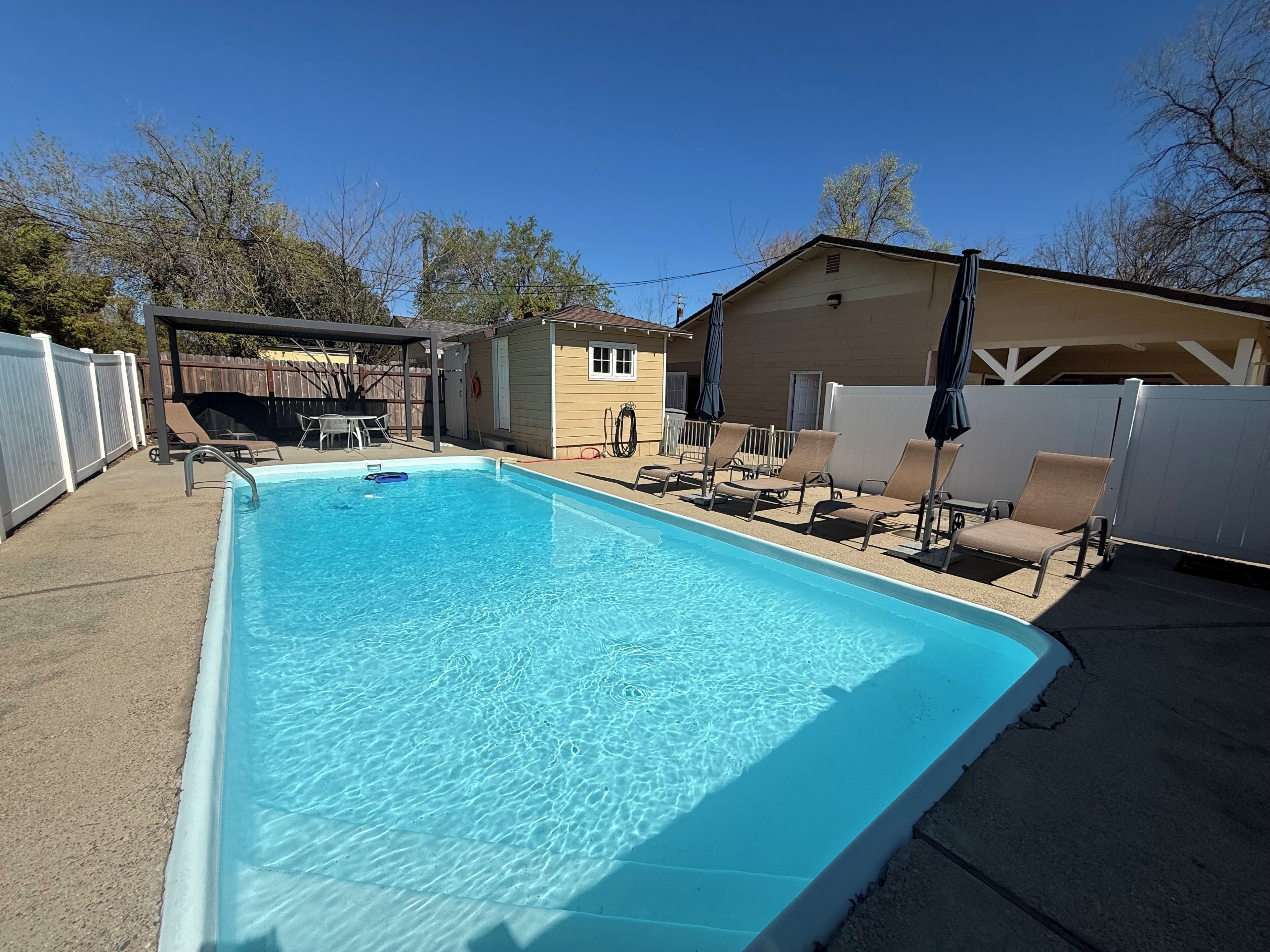 A rectangular swimming pool with clear blue water, surrounded by a white fence, and shaded by umbrellas.