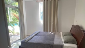 Une chambre à coucher avec un lit, une tête de lit en bois, une fenêtre avec des rideaux et une vue sur la verdure à l’extérieur.