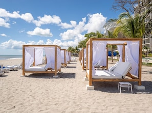Une plage avec des lits de repos surmontés de toiles en bois, des coussins blancs et un ciel d’un bleu limpide.