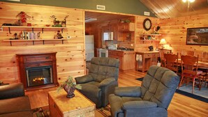 L’intérieur d’un chalet confortable en bois, avec une cheminée, deux fauteuils, un coin repas et une cuisine.