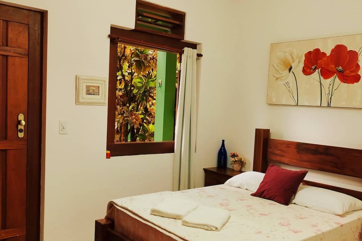 Quarto bem arrumado com cama de madeira, ventoinha no teto, janela com cortinas, pintura de flores e porta de madeira.