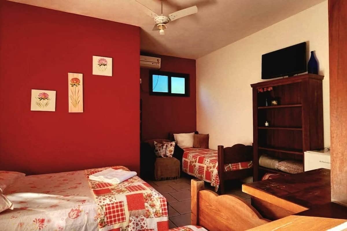 Quarto com duas camas, uma escrivaninha de madeira, uma estante e uma televisão sobre um suporte.