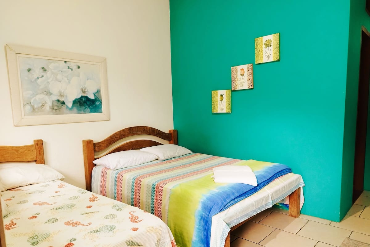 Quarto com duas camas de madeira, uma com colcha listrada e colorida e outra com colcha branca estampada. Há um quadro emoldurado na parede acima das camas, e três pequenos objetos decorativos na parede verde-água.