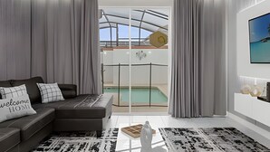 A modern living room with a sofa, a coffee table, and a view of a swimming pool through a glass door. - 4BR house at Storey Lake Resort 9min Disney (Kissimmee)