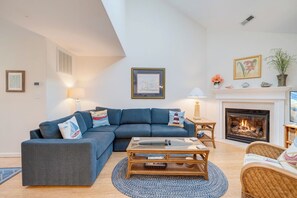 Ein Wohnzimmer mit einem blauen Sofa, einem Glastisch, einem Kamin und einem kleinen Beistelltisch mit einer Lampe.