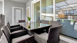 A modern dining area with a glass table and wicker chairs, large windows, and a view of a building across the street.