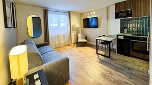 A modern living room with a sofa, a TV, a dining table, and a kitchen area with a microwave. - Cozy mountain studio | Summer pool + parking (Les Houches)