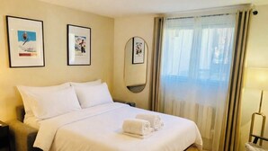 A hotel room with a bed, a bedside table, a chair, a window with curtains, and framed pictures on the wall. - Cozy mountain studio | Summer pool + parking (Les Houches)