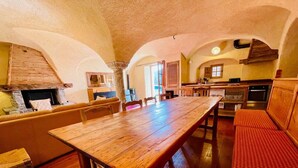 A cozy dining area with a wooden table and benches, a kitchen in the background, and a stone wall.