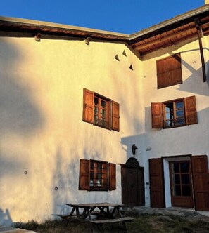 Un edificio con múltiples ventanas y puertas de madera, un patio empedrado con piedra y una mesa de picnic.