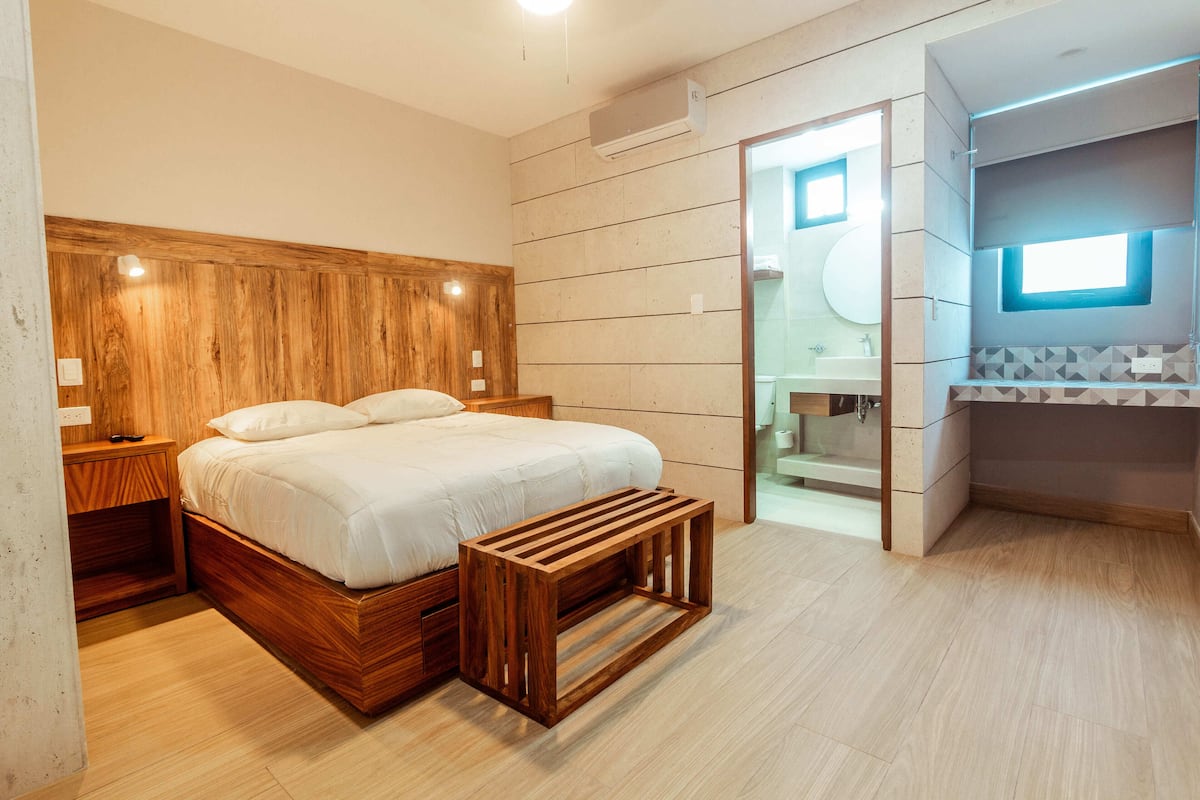 A bedroom with a wooden headboard, a bed with white bedding, a wooden bench, and a bathroom with a window and tiled walls.
