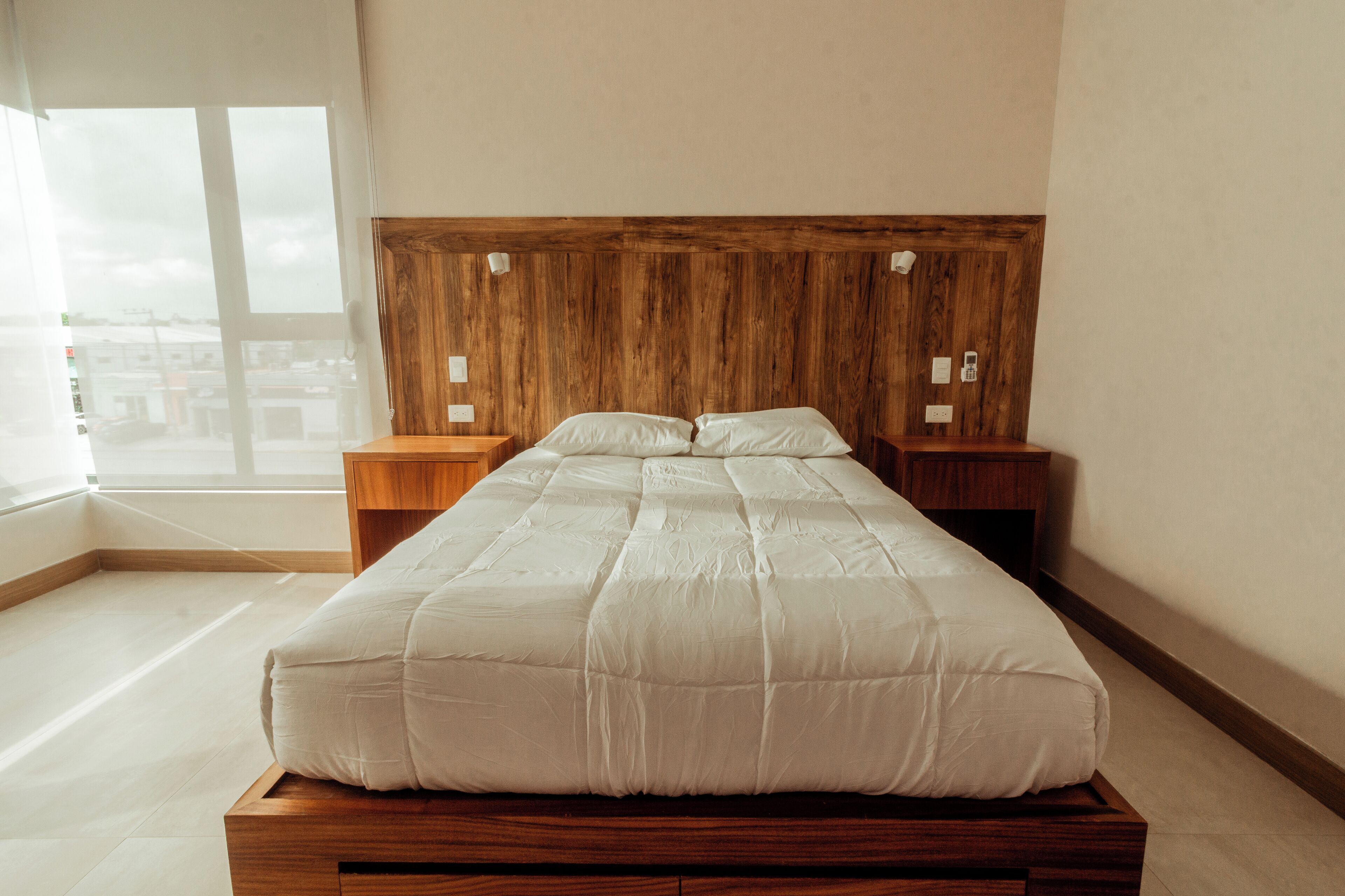 A bedroom with a wooden headboard, a large bed with white bedding, two bedside tables, and a large window providing a view of the city.
