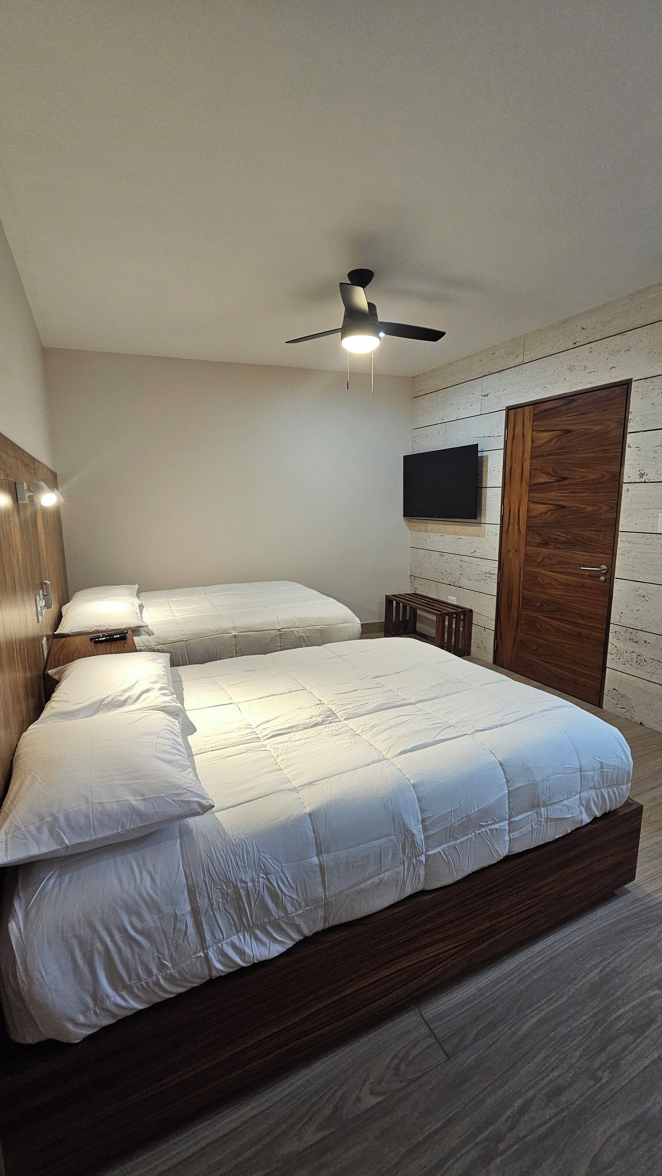 A bedroom with a large bed, a ceiling fan, a wooden wardrobe, and a mounted TV.