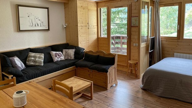 L’intérieur chaleureux d’un chalet avec un canapé en bois, une petite table, un lit et de grandes fenêtres donnant sur le paysage extérieur.