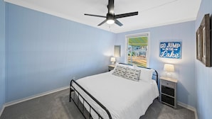 Quarto com cama, ventoinha no teto, mesa de cabeceira com abajur e um letreiro na parede.
