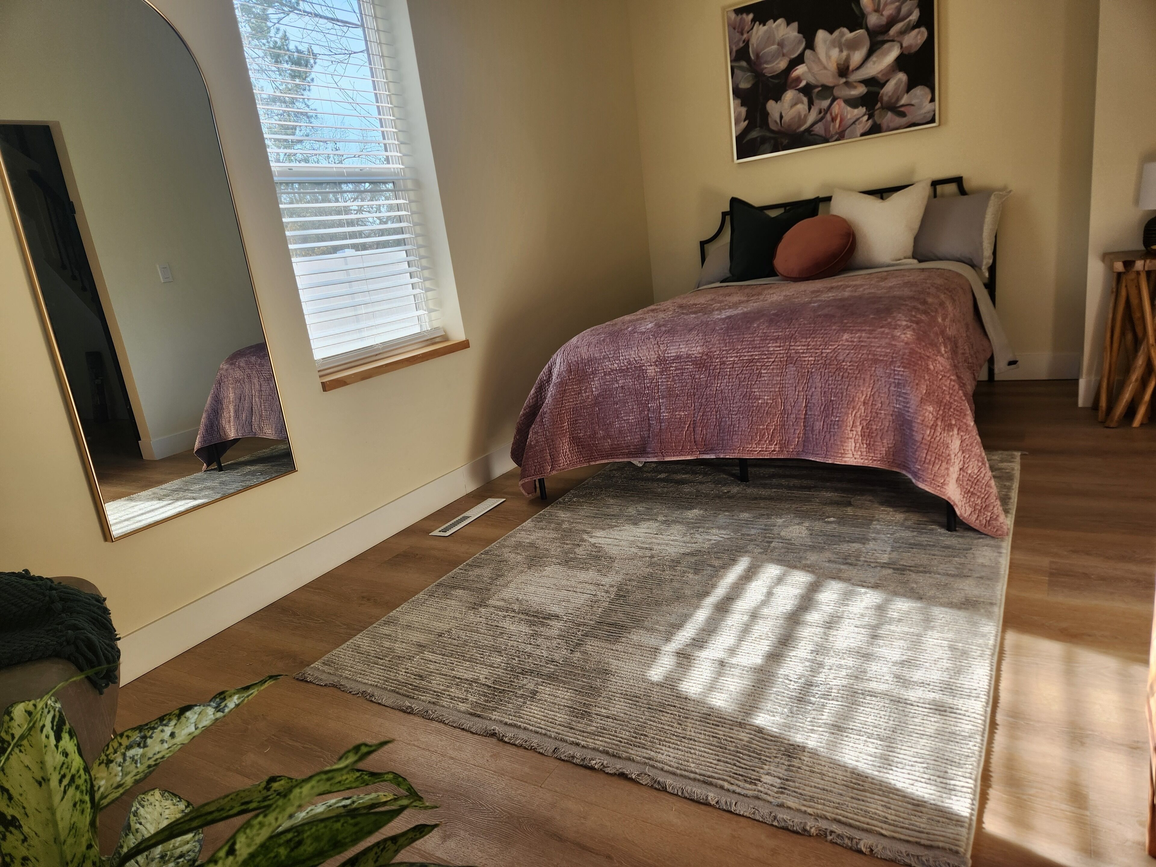 A bedroom with a bed, a large mirror, a window with blinds, a painting on the wall, and a plant.