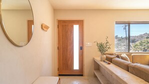 A modern living room with a wooden door, a large mirror, a sofa, and a view of a rocky landscape through a window. - Desert Nest: Cozy Retreat Amidst Boulders (Yucca Valley)