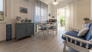 A cozy living room with a blue sofa, a dining table with chairs, a kitchen area with cabinets and a kettle, and a large window with curtains. - Casa Liliana Ac Wifi Forte Dei Marmi (Forte dei Marmi)