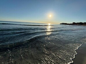 A beach with clear water reflecting the sun, a sandy shore, and a distant shoreline with buildings. - Mobile home with air conditioning, terrace and pool (Santa Lucia/Nuoro)