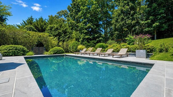 A rectangular swimming pool with clear blue water, surrounded by a paved deck. There are four lounge chairs aligned along the pool's edge.