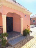 Un edificio con fachada rosa, puerta negra y columnas blancas. Hay escalones que conducen a la entrada, y un pequeño jardín con plantas y flores.