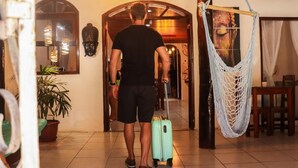 A person walking through a hallway with a suitcase, a hanging hammock, and a door with a decorative arch. - Pousada Pura Vida (Itacaré)