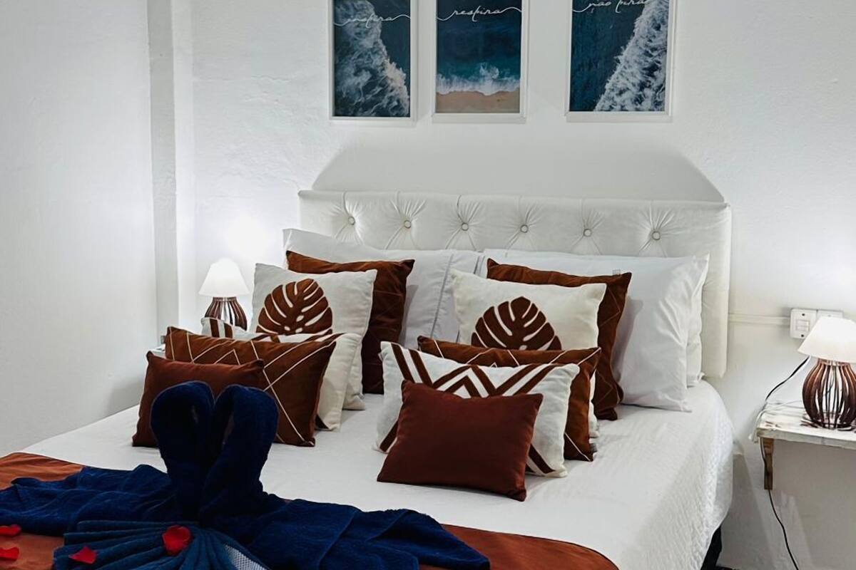 A neatly arranged bedroom with a large bed, multiple pillows, a bedside table with a lamp, and three framed pictures on the wall.