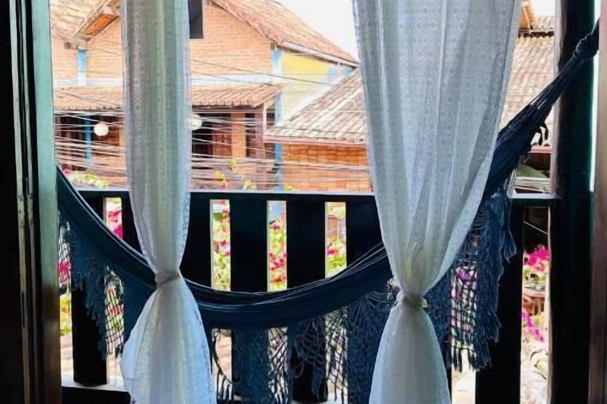 A room with a bed, a hammock, and a view of buildings through a window with sheer curtains.