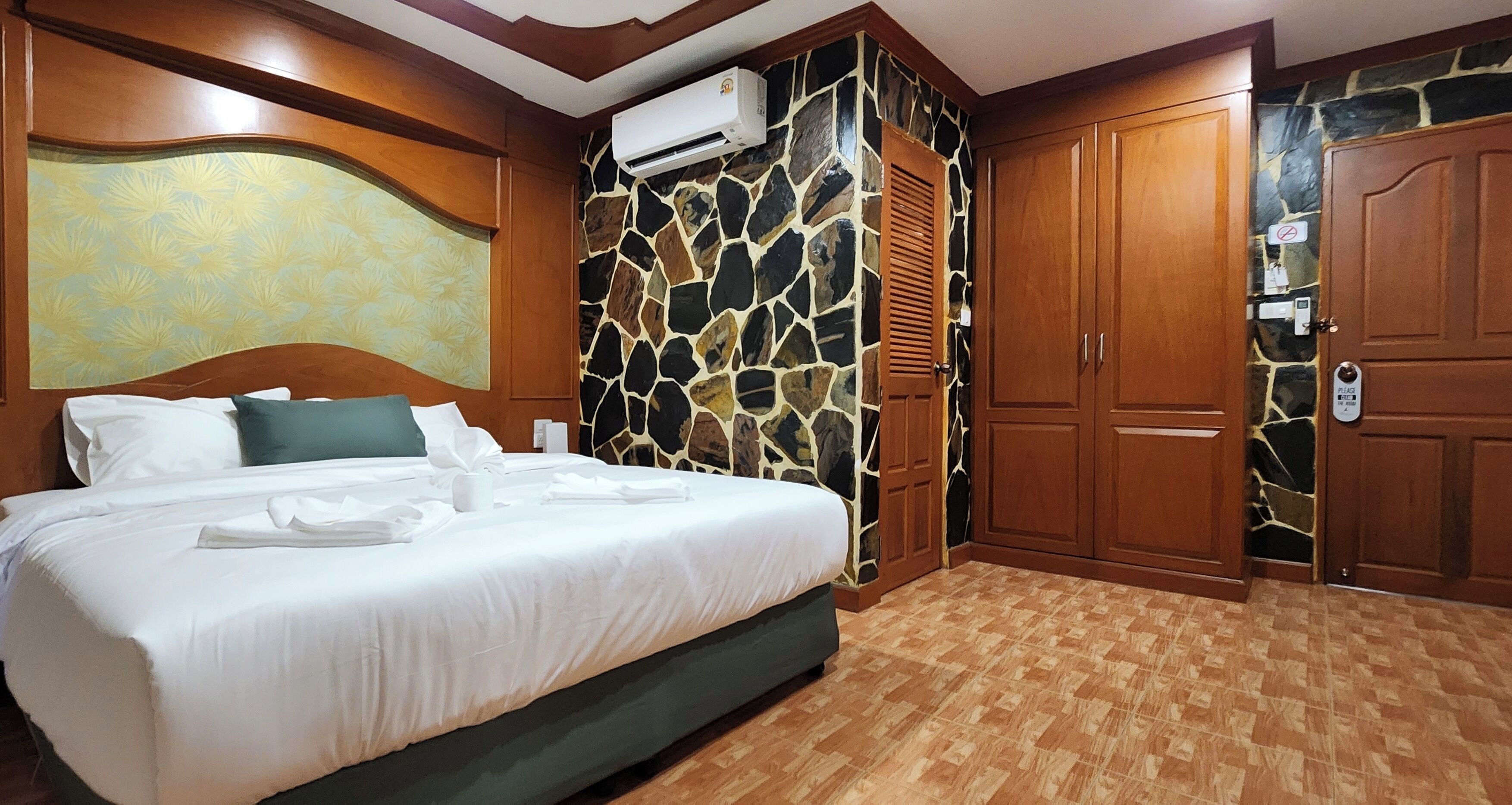 A hotel room with a large bed, a stone wall, wooden wardrobe, and an air conditioning unit.