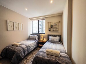 Un dormitorio con dos camas, una ventana, una mesita de noche con lámpara y decoración en la pared.