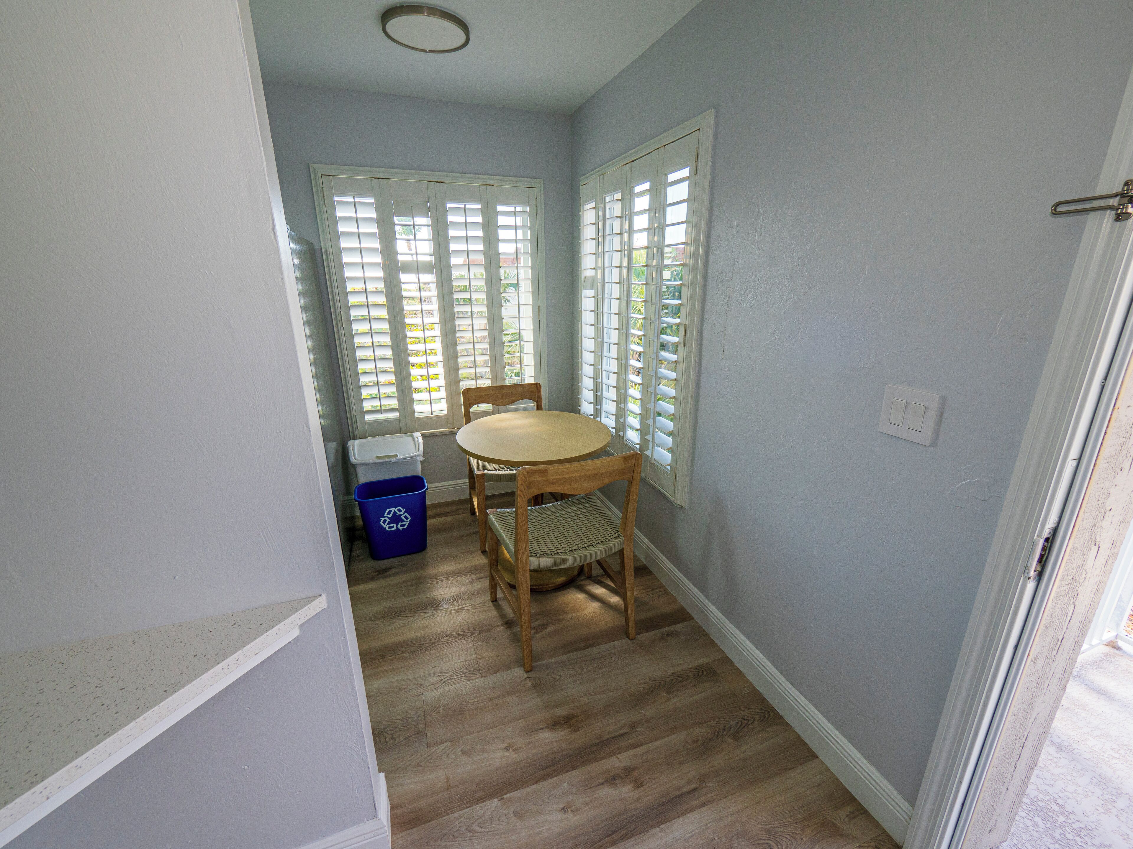 A room with a wooden floor, a round table, and two chairs. There are white-framed windows with blinds, a white door, and a blue recycling bin.
