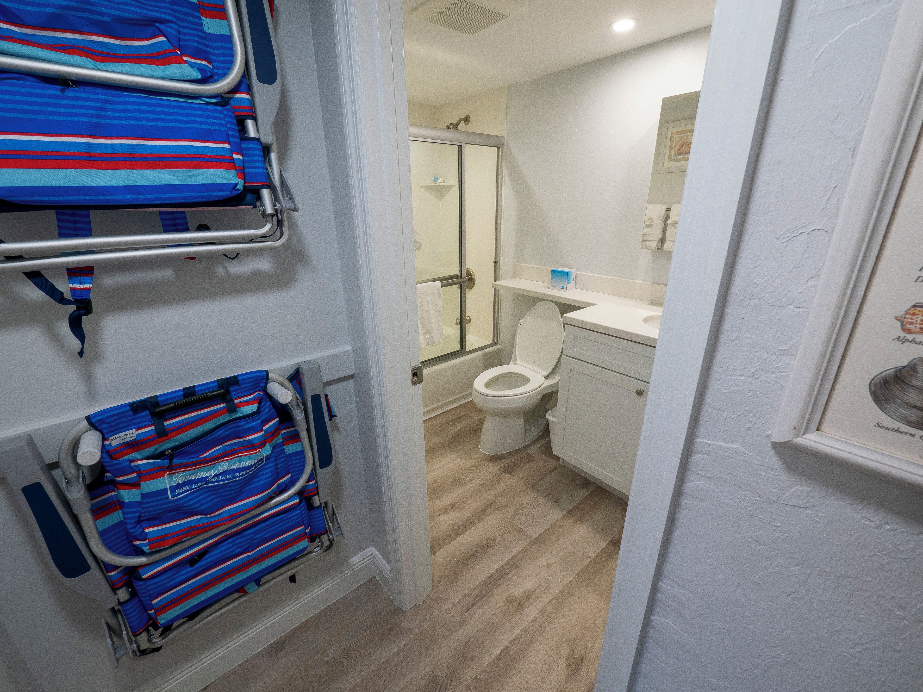 Un baño con inodoro blanco, lavamanos y ducha. Hay toallas azuladas a rayas y una silla de ruedas en un rincón.