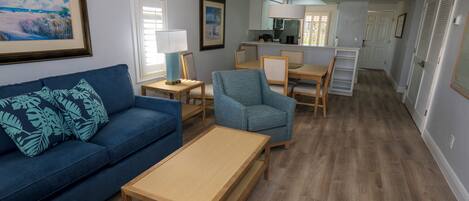 A living room with a blue sofa, a wooden coffee table, and a blue armchair. There is a dining area with a table and chairs, and a kitchen in the background.