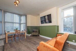 A living room with a wooden dining table, four chairs, a green accent wall, a large window, and an orange sofa. - Luxe Living (Huddersfield)