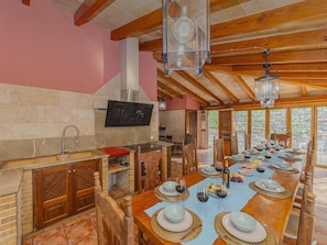 A dining area with a long table set for a meal, a kitchen with a modern stove, and a stone wall. - Villa Can Emmes · Villa with private pool and ample outdoor space in Alcúdia. (Alcúdia)