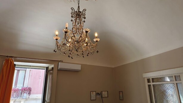 A room with a chandelier, a window with curtains, a flat-screen TV, and a wall-mounted air conditioner. - RETROETNEA - Retro-style Apartment in historic center with 2 connecting bedrooms (Catania)