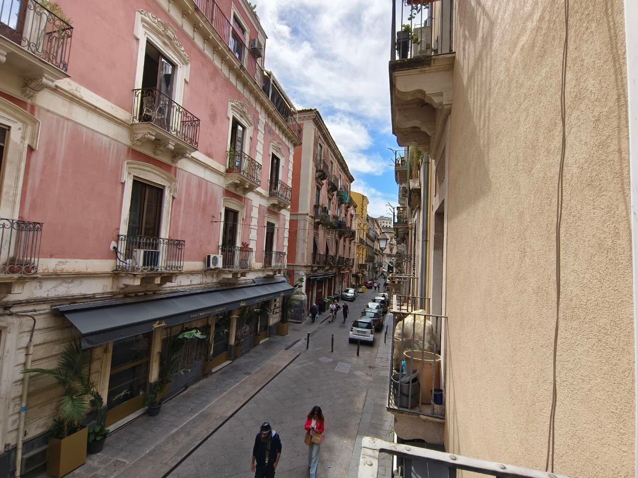 A narrow street with buildings on both sides, featuring balconies and a row of parked cars.