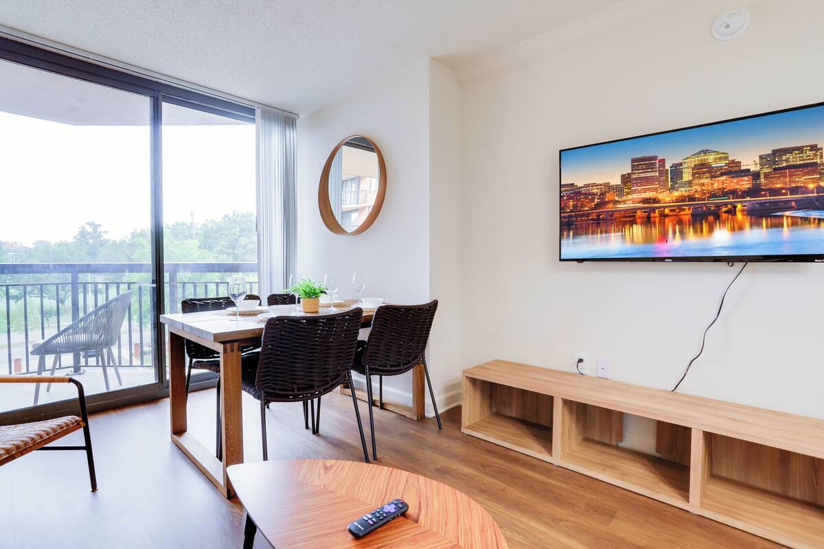 A modern living room with a large TV, a dining area with a table and chairs, and a balcony with a view.