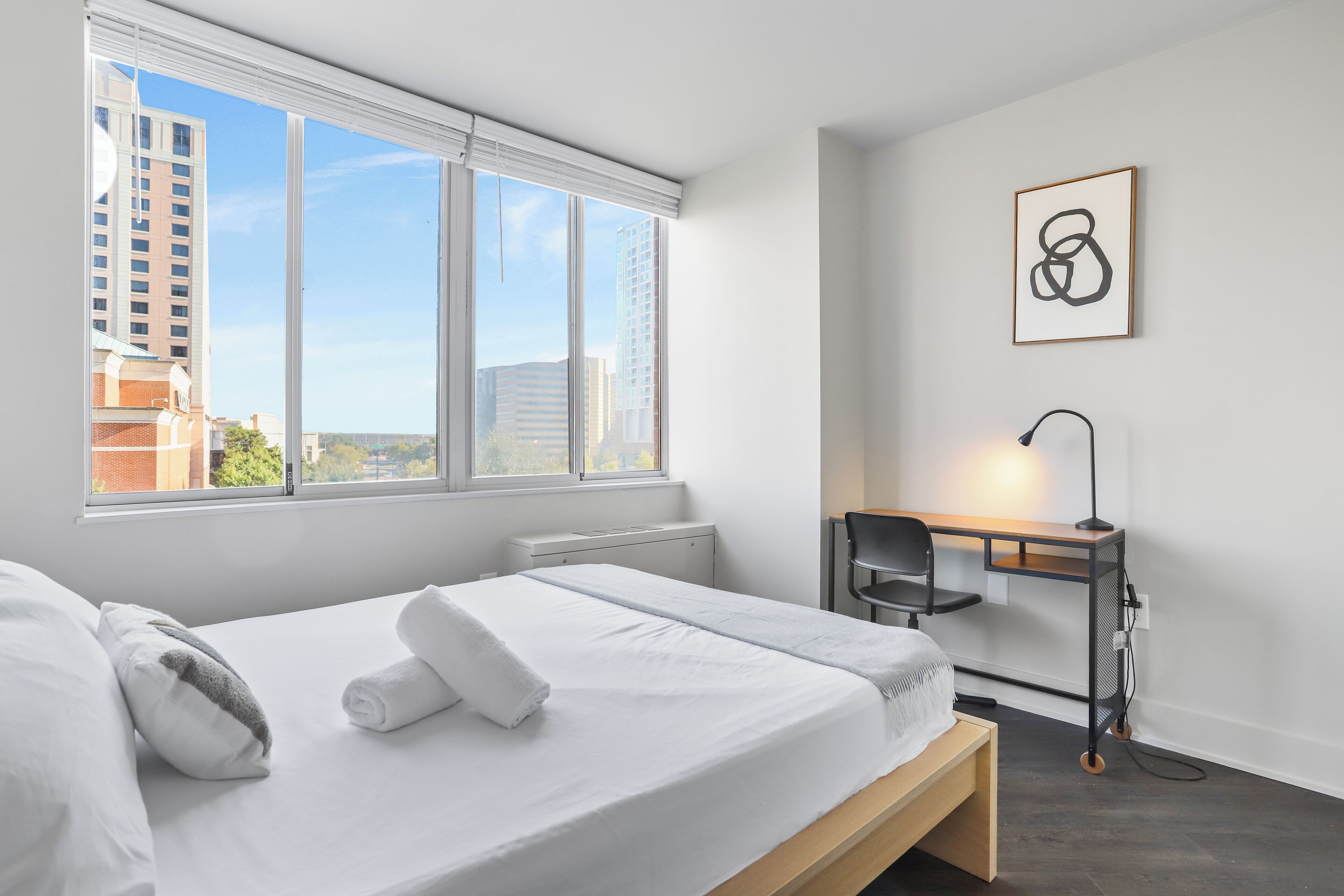 A hotel room with a bed, a desk, a chair, and a view of buildings through large windows.