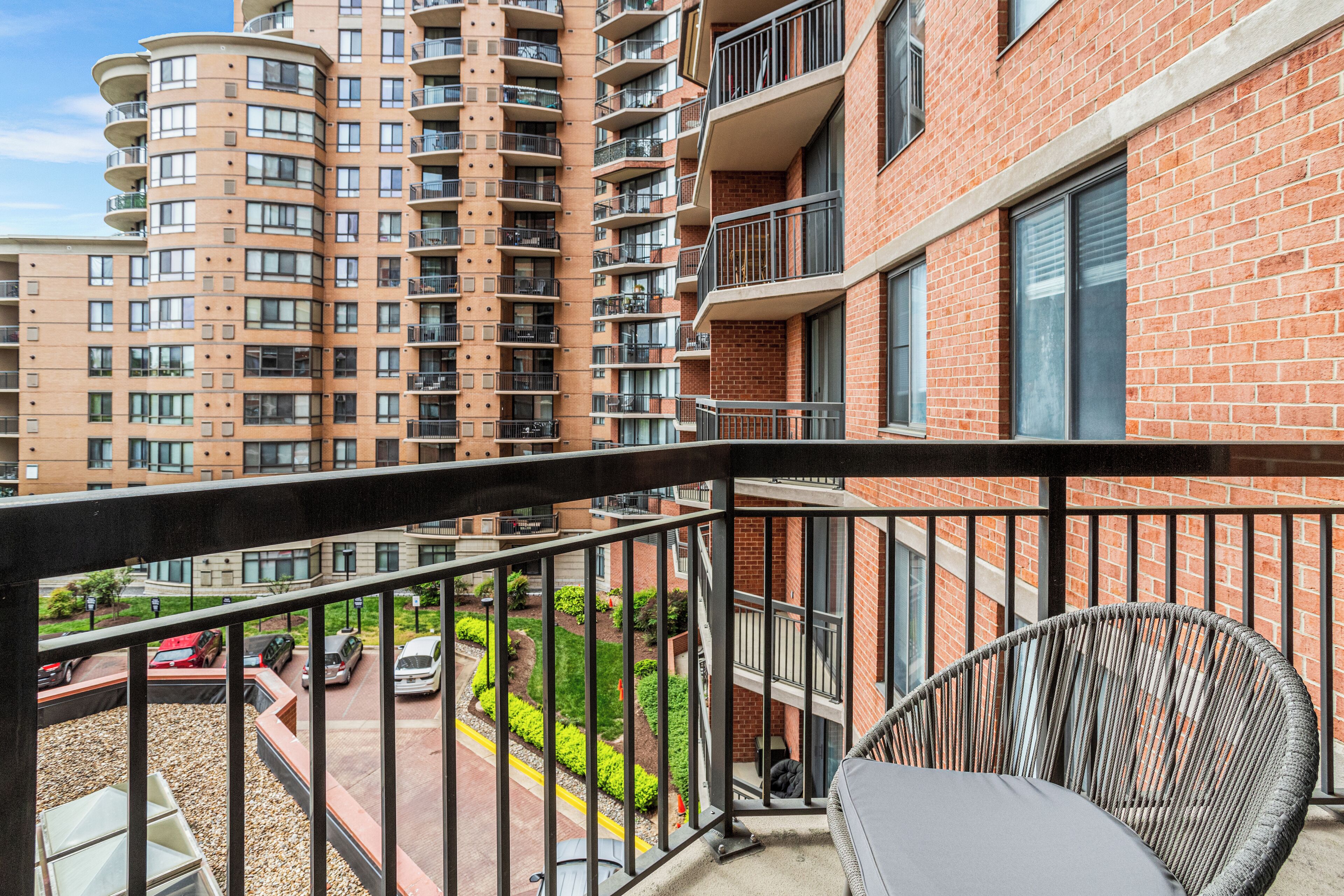 A balcony with a wicker chair and a view of a parking lot and adjacent buildings.