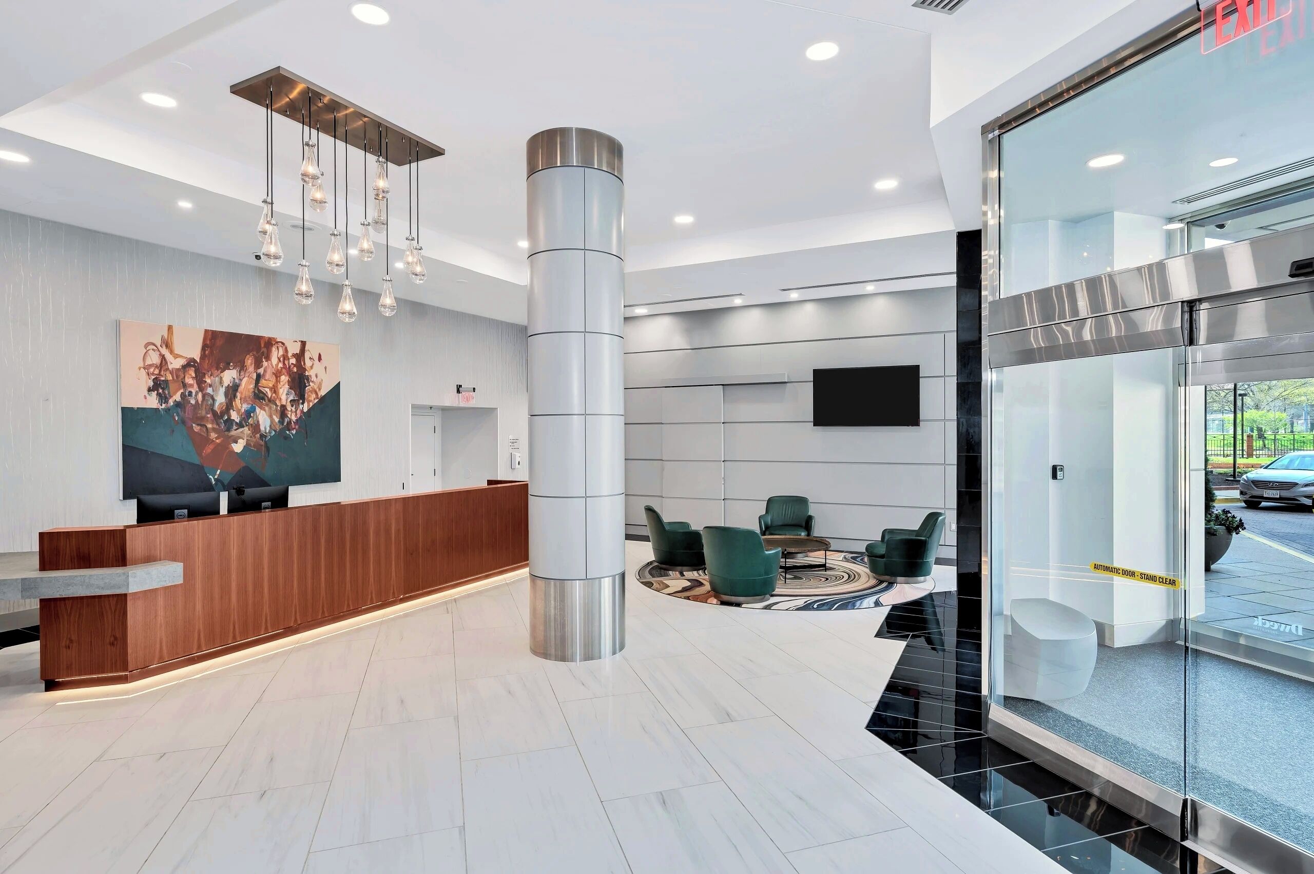 A modern reception area with a wooden reception desk, a large abstract painting, a central pillar, and a seating area with green chairs and a round table.
