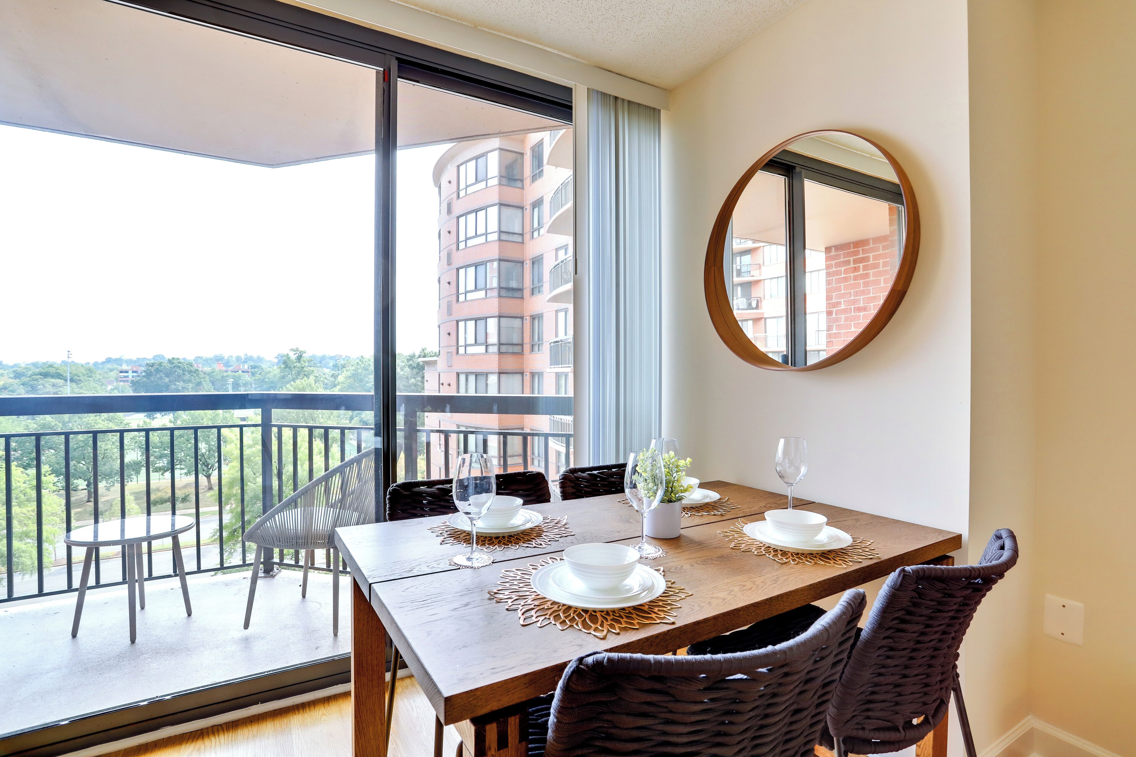 Un comedor con vista a un balcón, un espejo redondo y una mesa dispuesta con platos y vasos blancos.