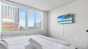 Une chambre d’hôtel dotée d’une grande fenêtre, d’un lit avec du linge de lit blanc, d’un téléviseur à écran plat fixé au mur et d’un tableau représentant une scène de plage.