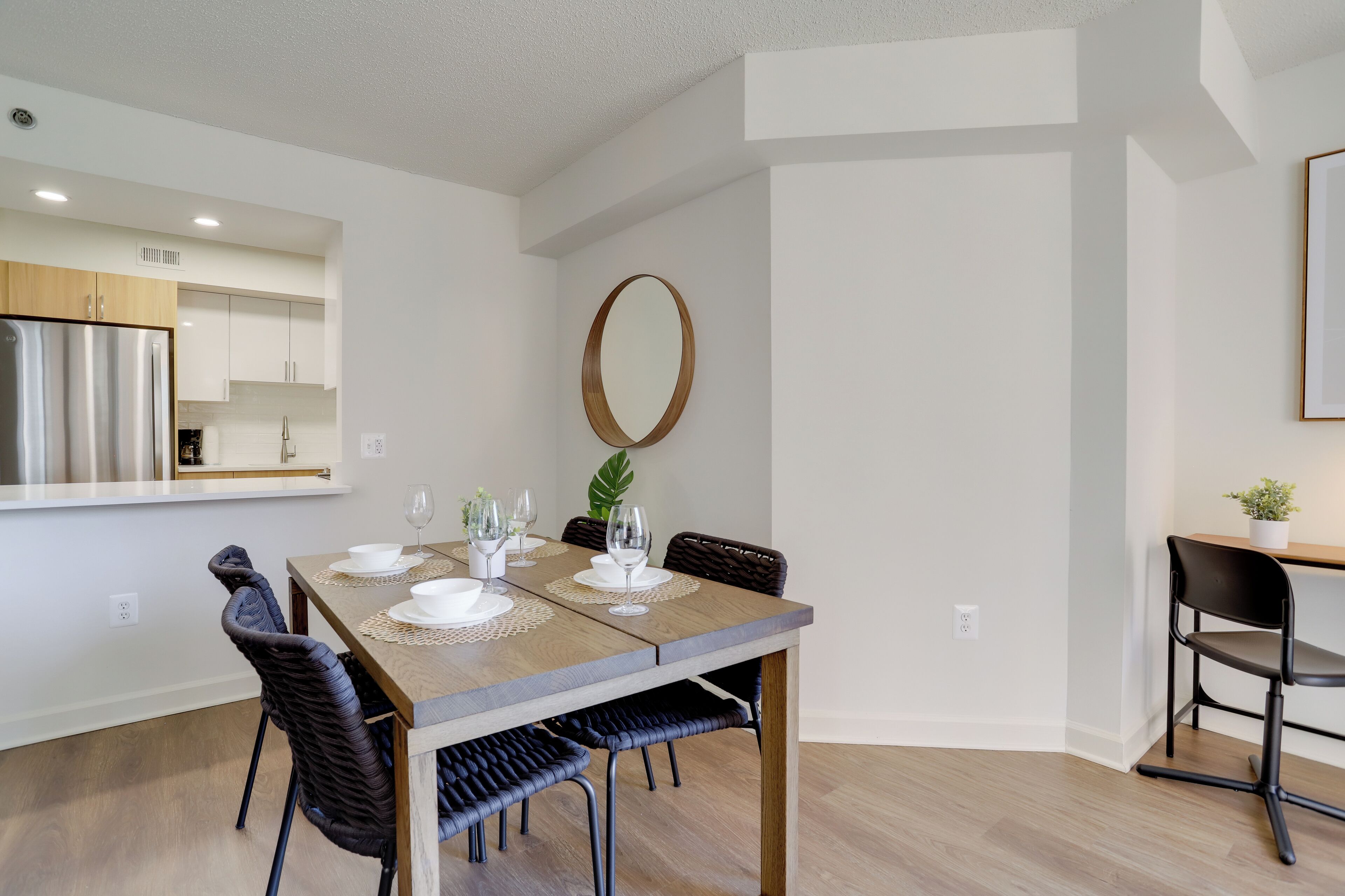 A dining area with a wooden table set for four, surrounded by dark chairs, a mirror on the wall, and a kitchen in the background.