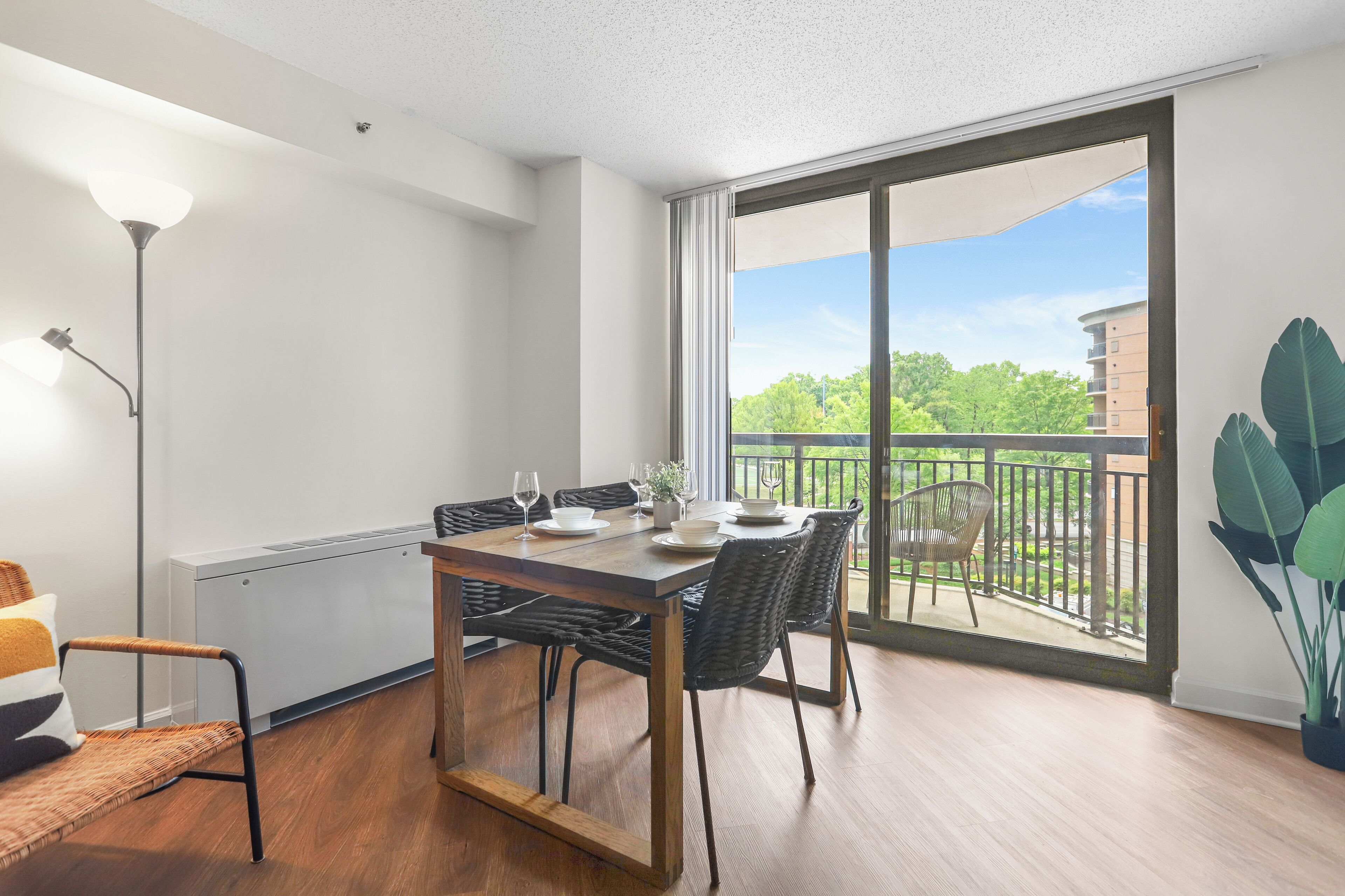 Ein moderner Essbereich mit einem Holztisch, schwarzen Korbstühlen und Blick auf einen Balkon mit Grünpflanzen und Gebäuden im Hintergrund.